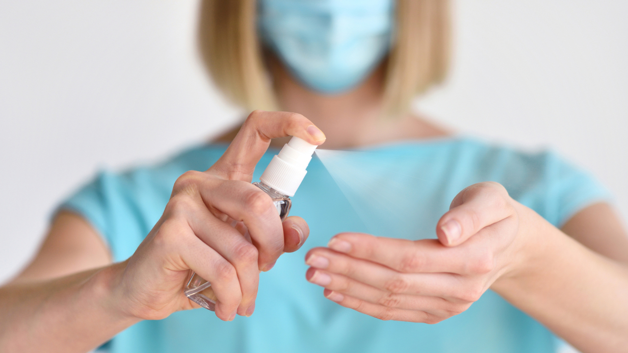 yüzünde cerrahi maske bulunan bir kadın eline dezenfektan sıkarken görülüyor
