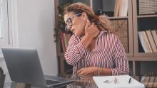 Bir kadın, boyun ağrısı çeken bir şekilde bir masa başında bir dizüstü bilgisayarın başında oturuyor.